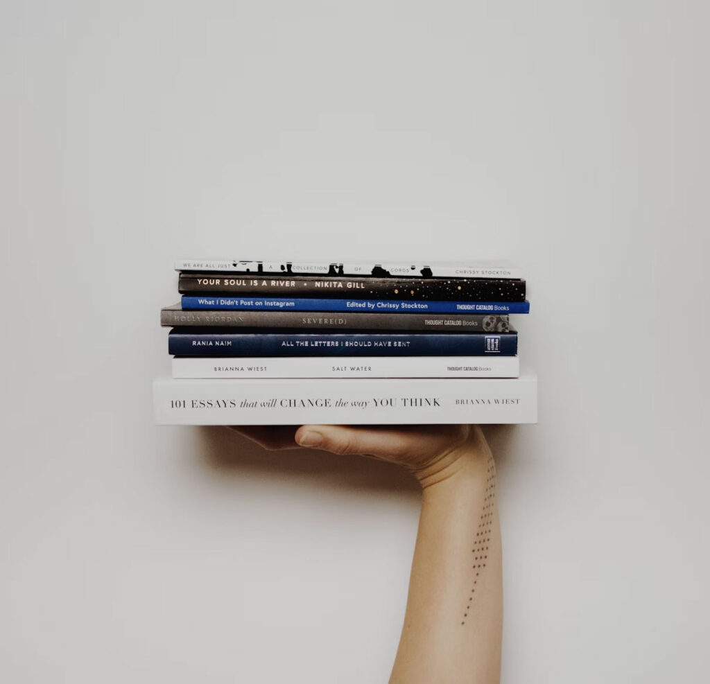 A hand holding a stack of books with titles like "Your Soul is a River" and "101 Essays That Will Change The Way You Think.